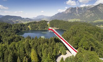 An impressive viewpoint bridge over a clear lake, surrounded by green forests and mountains. The sky is blue with some clouds.