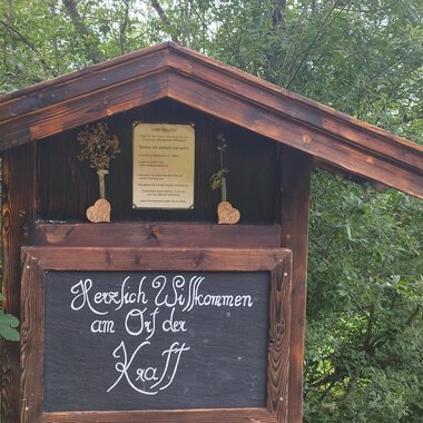 A traditional wooden sign with the inscription "Warm Welcome to the Ötztal". Surrounded by green trees and a rural setting. | © Nicole Lughammer