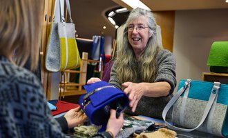 Eine Verkäuferin lächelt und zeigt eine blaue Tasche einer Kundin. Im Hintergrund sind weitere Taschen und Handarbeiten sichtbar.