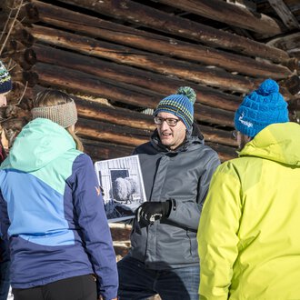 A group of five people stands in the snow in front of a wooden barn. They are collectively looking at a photo that one of the participants is holding in their hand. | © Kleinwalsertal Tourismus | Dominik Berchtold