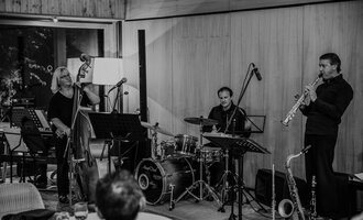 Three musicians are happily gathered together, playing various instruments. In the background, the nature hotel can be seen.