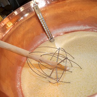 In einem großen Kupferkessel wird eine Mischung zubereitet. Ein Holzrührer wird in die goldene Masse eingetaucht.