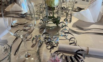 Two champagne glasses toast on a festively decorated table. In the background, red Christmas ornaments and golden decorations can be seen. | © Genuss- & Aktivhotel Sonnenburg