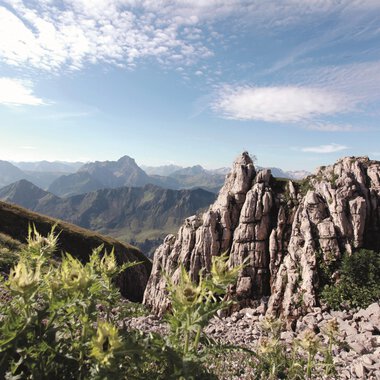 Eine beeindruckende Berglandschaft mit schroffen Felsen und majestätischen Bergen im Hintergrund. Der Himmel ist klar und die Umgebung ist von grünem Pflanzenbewuchs geprägt. | © Kleinwalsertal Tourismus eGen | Frank Drechsel