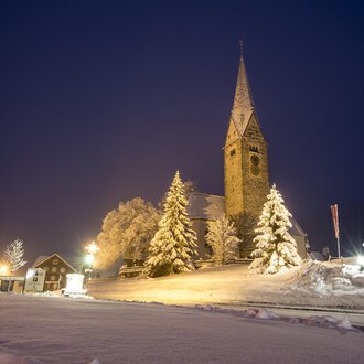 A snowy landscape at night with a church and illuminated trees. The sky is dark blue and the road is visible. | © Kleinwalsertal Tourismus | Dominik Berchtold