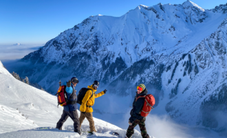 Drei Personen in winterlicher Ausrüstung stehen auf einem schneebedeckten Berg. Im Hintergrund erstrecken sich beeindruckende Berge unter einem klaren blauen Himmel.
