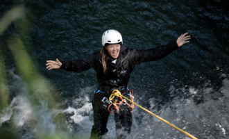 A person enthusiastically climbs on a rope over water. They are wearing a helmet and are dressed in a protective suit.