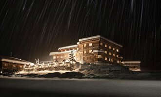 A cozy wooden building in the snow, illuminated by warm light. In the background, gentle snowflakes fall through the night. | © Auenhütte | Yorck Dertinger