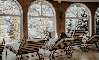 Ein gemütlicher Raum mit Liegen und großen Fenstern. Draußen ist eine winterliche Landschaft mit Schnee und Bäumen zu sehen. | © Verwöhn- & Wellnesshotel Walserhof| Aileen Melucci