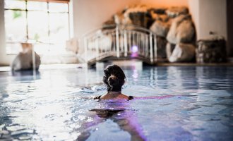 Eine Frau schwimmt in einem entspannenden Innenpool. Im Hintergrund sind Felsen und sanfte Beleuchtung zu sehen. | © Verwöhn- & Wellnesshotel Walserhof| Aileen Melucci