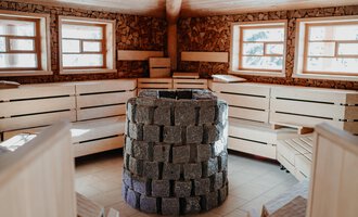 A cozy sauna room with wooden benches and large windows. In the center stands a stone oven. | © Verwöhn- & Wellnesshotel Walserhof| Aileen Melucci