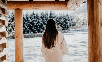 A woman in a white bathrobe stands under a wooden canopy and looks at a snowy landscape. The trees in the background are covered in snow. | © Verwöhn- & Wellnesshotel Walserhof| Aileen Melucci