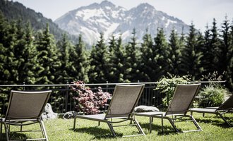 A peaceful terrace with deck chairs and a view of majestic mountains. In the background, there are green trees and colorful plants. | © Verwöhn- & Wellnesshotel Walserhof| Aileen Melucci