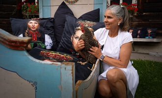 A woman is sitting on a colorful couch and holding a chicken in her arms. In the background, artistically designed cushions can be seen. | © Annett Schwärzler Nails & Spa | Stefanie Haid