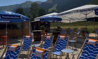 A beach area with blue loungers and umbrellas. In the background, green mountains can be seen. | © MORE VIBES by Alex Lambsdorff