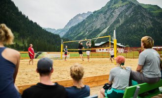 Ein Beachvolleyballspiel in den Bergen mit einer Gruppe von Zuschauern. Im Hintergrund sind grüne Wälder und majestätische Berge zu sehen. | © Kleinwalsertal Tourismus | Oliver Farys