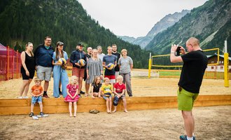 A group of people is standing on a sandy court with a volleyball net in the mountains. Someone is taking photos of them as they smile and pose. | © Kleinwalsertal Tourismus | Oliver Farys