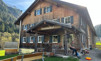 A rustic wooden house named "Gemstelhof" with an inviting porch. In the foreground, you can see a person sitting on a bench, surrounded by green landscape and mountains. | © Bernhards Gemstel Hofladen | Bernhard Heim
