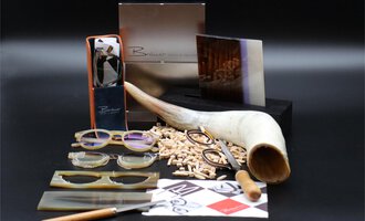 A stylish arrangement of glasses, wooden pieces, and tools. In the background, elegant packaging and a large horn bowl can be seen. | © Bräuer Optik & Akustik | AlpenMedia-Design