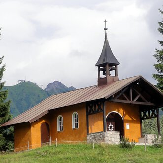 A small, orange church stands surrounded by green trees. In the background, mountains and a cloudy sky can be seen. | © Bruder Klaus Kapelle | Monika Gmeiner