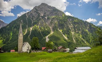 Eine malerische Landschaft mit einem Dorf und einer beeindruckenden Bergkulisse. Im Vordergrund steht eine alte Kirche und saftige Wiesen. | © Kleinwalsertal Tourismus | Steffen Berschin