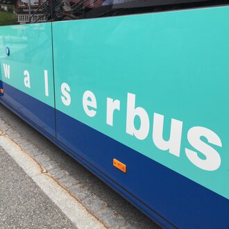 A bus with the inscription "walserbus" in teal and blue. The bus is at a stop. | © Kleinwalsertal Tourismus
