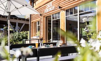 An inviting café with a wooden facade and a sunny outdoor area. Tables are set up for guests to enjoy the fresh air.