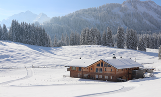 A beautiful wooden house in a snow-covered landscape. In the background, tall mountains and lush fir trees stand.