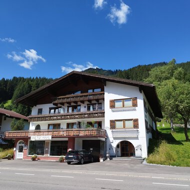 A charming building in alpine style with various balconies. In the background, green hills and a blue sky can be seen. | © Kleinwalsertal Tourismus | Nicole Lughammer