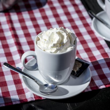 A cup with cream and a spoon is on a table with a red-and-white checkered tablecloth. In the background, more cups can be seen. | © Kleinwalsertal Tourismus | Dominik Berchtold