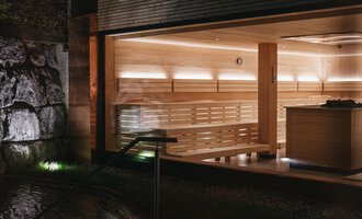 Eine moderne Sauna mit hellem Holzinterieur und sanfter Beleuchtung. Im Hintergrund sind Steine und eine entspannende Atmosphäre zu sehen. | © Naturhotel Chesa Valisa