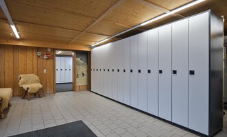 Ein moderner Umkleideraum mit weißen Spinden an einer Wand. Der Raum hat eine Holzdecke und Fliesenboden. | © Sport Kessler GmbH