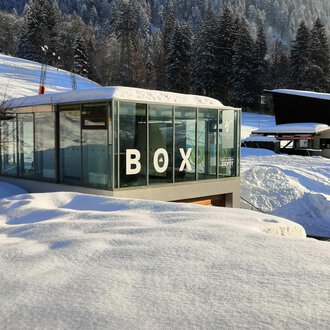 A modern glass building with the inscription "BOX" stands amidst a snow-covered landscape. In the background, snow-covered trees and additional buildings can be seen. | © Sport Kessler GmbH