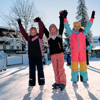 Eine Gruppe fröhlicher Kinder steht auf einer Eislauffläche und hebt die Hände in die Luft. Sie tragen bunte Winterkleidung und genießen den sonnigen Wintertag. | © Kleinwalsertal Tourismus | Oliver Farys