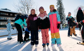 Three girls are standing on an ice rink, surrounded by other children who are ice skating. It is a sunny day with a clear sky and snow in the surroundings. | © Kleinwalsertal Tourismus | Oliver Farys