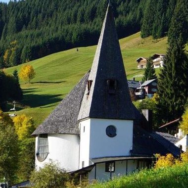 Eine malerische Kirche mit einem hohen spitzen Turm steht in einer grünen Landschaft. Im Hintergrund sind sanfte Hügel und einige Häuser zu sehen. | © Ev. Kirche Kleinwalsertal
