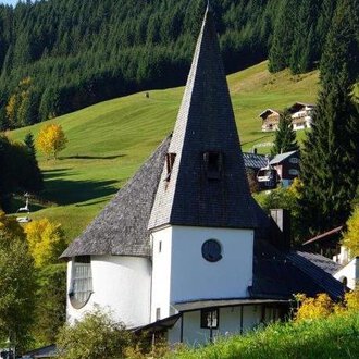 A picturesque church with a tall spire stands in a green landscape. In the background, there are gentle hills and some houses. | © Ev. Kirche Kleinwalsertal