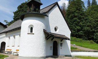 A small, white church with a distinctive roof and a bell tower. It is situated in a green surroundings with trees and a clear sky. | © Kleinwalsertal Tourismus | N. Lughammer