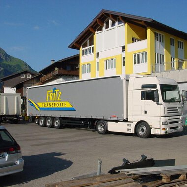 A truck from Fritz Transporte is parked in front of a large building. In the background, green mountains can be seen. | © Getränke Service Fritz | Helmut Fritz