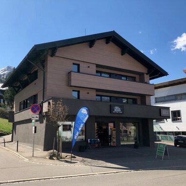 Ein modernes Gebäude an einer Straßenecke mit einem Geschäft im Erdgeschoss. Im Hintergrund sind Berge und andere Gebäude zu sehen. | © Sport Hilbrand GmbH