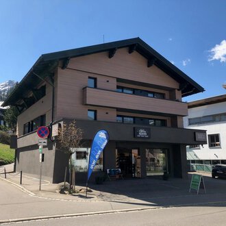 A modern building at a street corner with a shop on the ground floor. In the background, mountains and other buildings can be seen. | © Sport Hilbrand GmbH