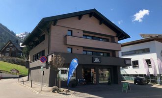 A modern building at a street corner with a shop on the ground floor. In the background, mountains and other buildings can be seen. | © Sport Hilbrand GmbH