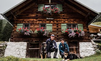 Ein rustikales Holzhaus mit blühenden Blumenkästen. Ein Paar mit zwei Hunden posiert davor. | © Hundewelt Kleinwalsertal | Stefan Lindbauer