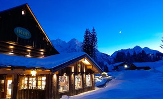 Der gemütliche iKuh-Laden in einem alten Bauernhaus im Schnee unter einem klaren, blauen Himmel. Die Berge im Hintergrund sind sanft beleuchtet, während der Abend naht.