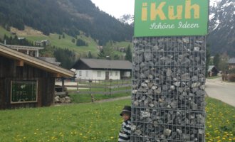 A child is standing next to a large sign that reads "iKuh" in the midst of a rural landscape. In the background, meadows and mountains can be seen. | © iKuh Schöne Ideen | Daniela Neumann
