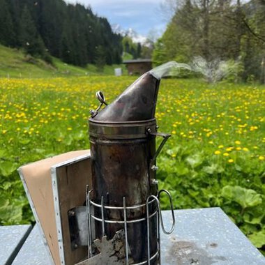 A smoking device is on a table in a blooming meadow. In the background, forests and a blue sky can be seen. | © Imkerei Jürgen Scharnagl