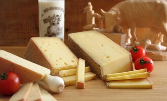 A piece of cheese lies on a wooden board, surrounded by fresh tomatoes and strips of cheese. In the background, there is a glass of milk and a wooden figure of an animal. | © Innere Wiesalpe | Andrea Kainz zuckerschnecke.at