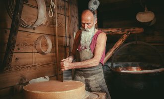 An older man with long white hair and a beard works in a woodworking shop. In the background, a large kettle can be seen. | © Käsestadl Kleinwalsertal | Nadine Paul