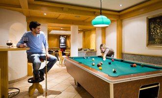 Eine gemütliche Spielhalle mit einem Billardtisch und einem Mann, der entspannt zuschaut. Eine Frau spielt konzentriert am Tisch. | © Aparthotel Kleinwalsertal Haller GmbH & Co KG