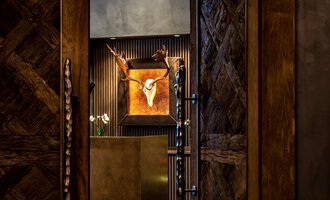 Ein stilvoller Eingangsbereich mit Holzwänden und einem dekorativen Geweih. Im Hintergrund ist ein Kunstwerk mit einem Tierkopf zu sehen. | © Travel Charme Ifen Hotel